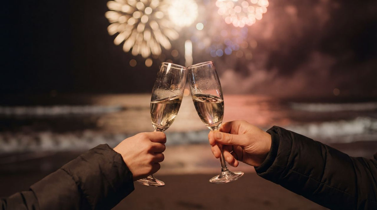 Two arms are toasting glasses of champagne while fireworks explode in the background