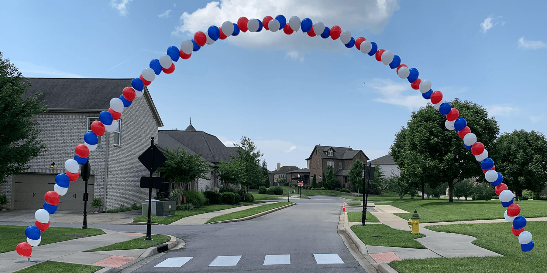 Balloon arch in red, white, and blue over a residential street with houses and trees in the background.